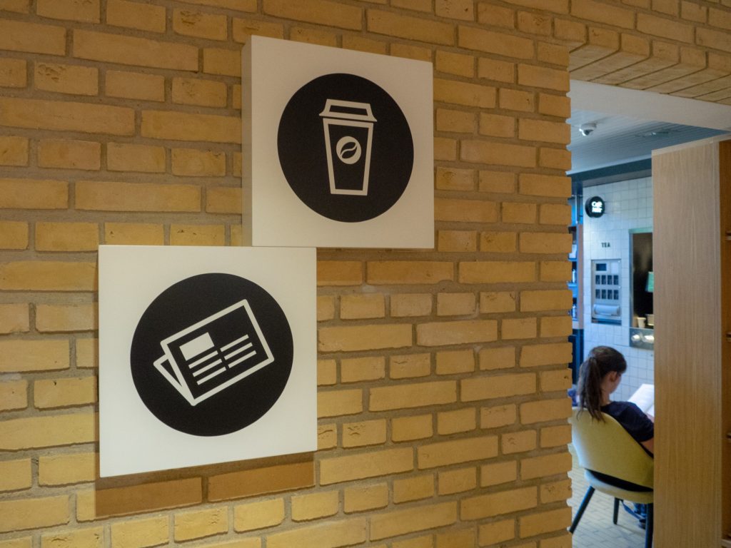 To hvide skiltekasser med piktogrammer for kaffebar og læsestof monteret på gul teglstensvæg; kaffebaren ses til højre.