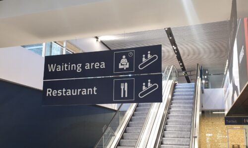 Wayfinding-skilt i lufthavn med piktogrammer og tekst der viser op ad rulletrappen mod restaurant og venteområde