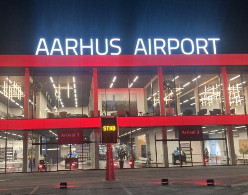 Stort oplyst velkomstskilt på taget af terminalbygning med rødt lys på facaden, set fra landingsområdet